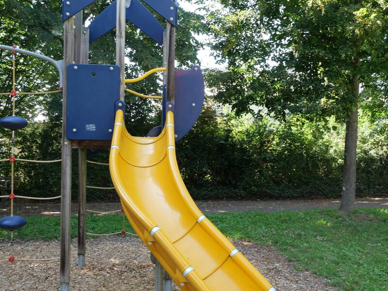Ein Spielplatzgerät mit einer gelben Rutsche vor einem Hintergrund aus grünen Bäumen.