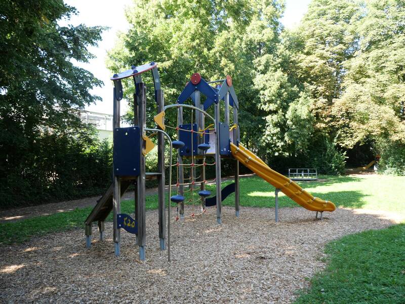 Ein Spielplatz mit einem Klettergerüst und einer gelben Rutsche steht auf Holzschnitzeln, umgeben von grünen Bäumen.