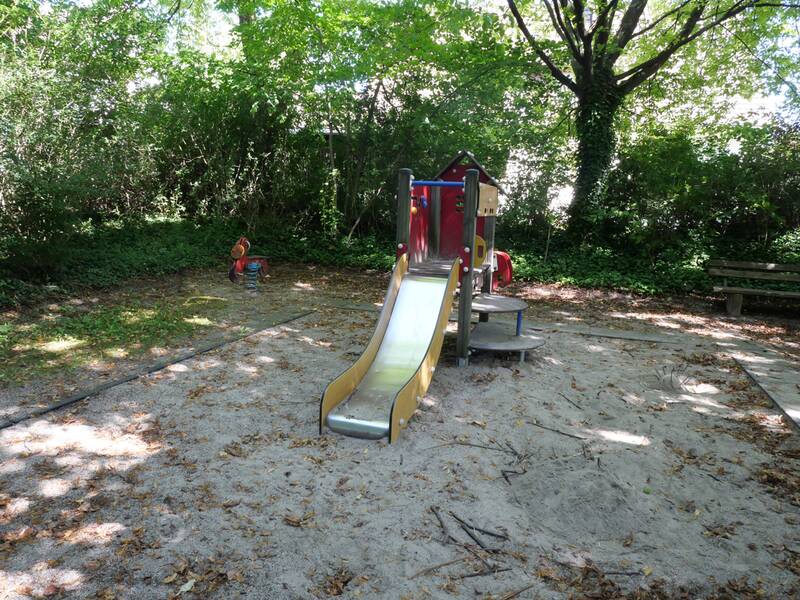 Ein kleiner Spielplatz mit einer Rutsche und einer Wippe, umgeben von Bäumen und Büschen.