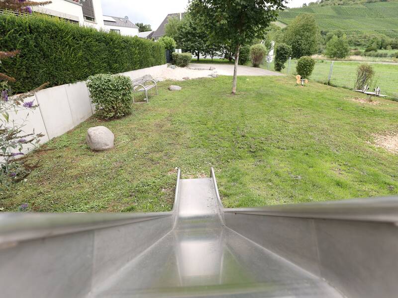 Blick von oben auf eine Rutsche, die auf eine grüne Rasenfläche zeigt, mit Sandfläche und Bank im Hintergrund.
