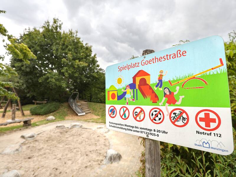 Ein Schild mit Regeln für den Spielplatz Goethestraße vor Sandkasten und Rutsche.