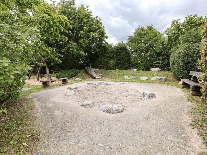 Ein runder Sandkasten mit großen Steinen am Rand, daneben Sitzbänke und im Hintergrund eine Rutsche.