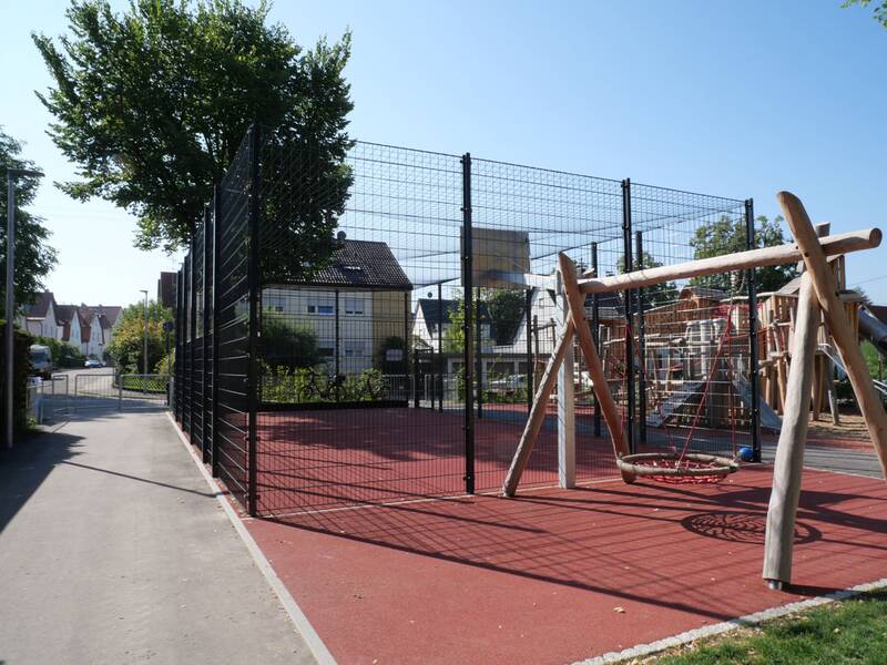 Ein eingezäunter Spielplatz mit einem Klettergerüst und einer Schaukel auf einem roten Bodenbelag, umgeben von Bäumen und einem Gehweg, bei sonnigem Wetter.