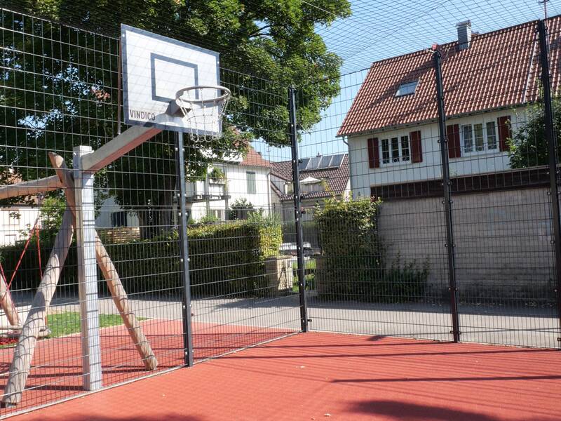 Ein Basketballplatz mit rotem Bodenbelag, umgeben von einem hohen Zaun, befindet sich inmitten eines Wohngebiets mit Bäumen und Häusern im Hintergrund.