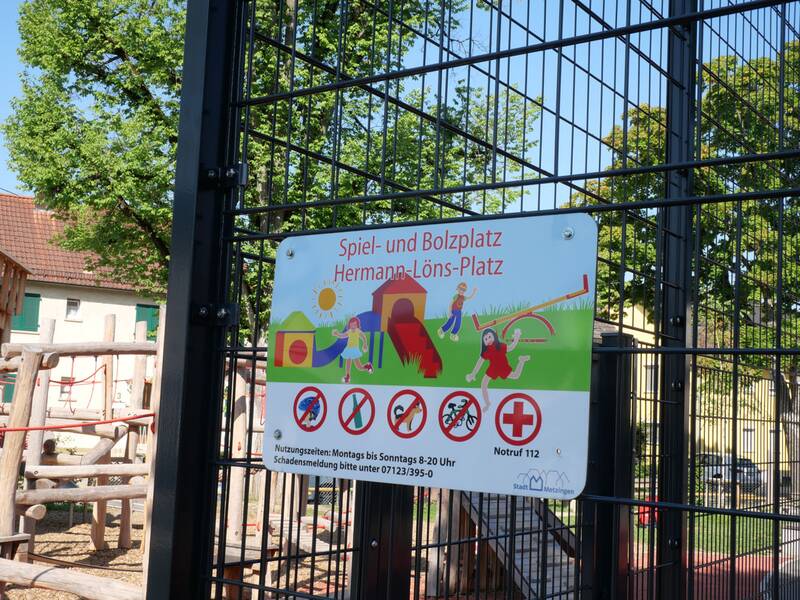 Ein Schild an einem Zaun weist auf den "Spiel- und Bolzplatz Hermann-Löns-Platz" hin und verbietet Hunde, Glasflaschen sowie Rauchen, während im Hintergrund Teile eines Spielplatzes zu sehen sind.
