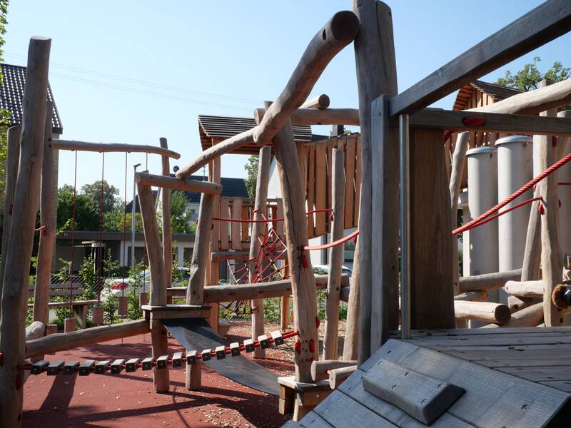 Ein moderner Spielplatz aus Holz mit verschiedenen Kletterelementen und roten Seilen befindet sich im Freien.