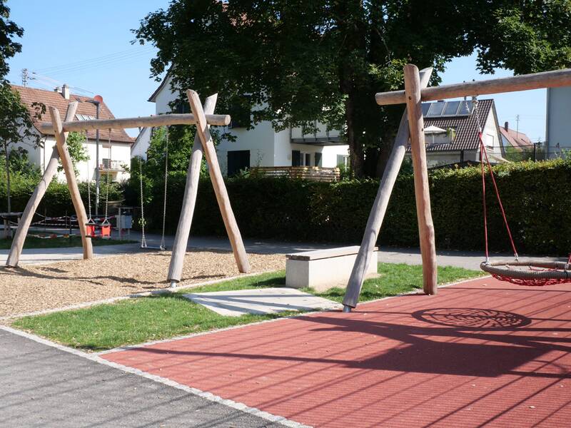 Ein Spielplatz mit Holzschaukeln, darunter eine Nestschaukel, umgeben von Häusern und Bäumen im Hintergrund.