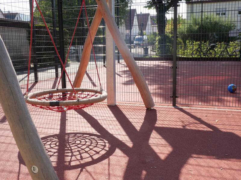 Ein Spielplatz mit einer runden Nestschaukel aus Seilen und einem blauen Ball auf dem Boden, umgeben von einem Gitterzaun.