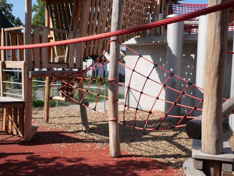 Ein Spielplatzklettergerüst aus Holz und roten Seilen mit einem Netz zum Klettern und einer Holzplattform im Hintergrund.
