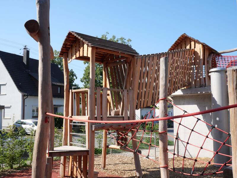 Ein hölzernes Klettergerüst mit rutschfestem Bodenbelag und einem Netz aus roten Seilen steht auf einem Spielplatz vor einem Haus.