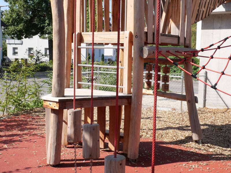 Ein hölzernes Klettergerüst mit roten Seilen und Balancierstämmen steht auf einem Spielplatz mit weichem Bodenbelag.
