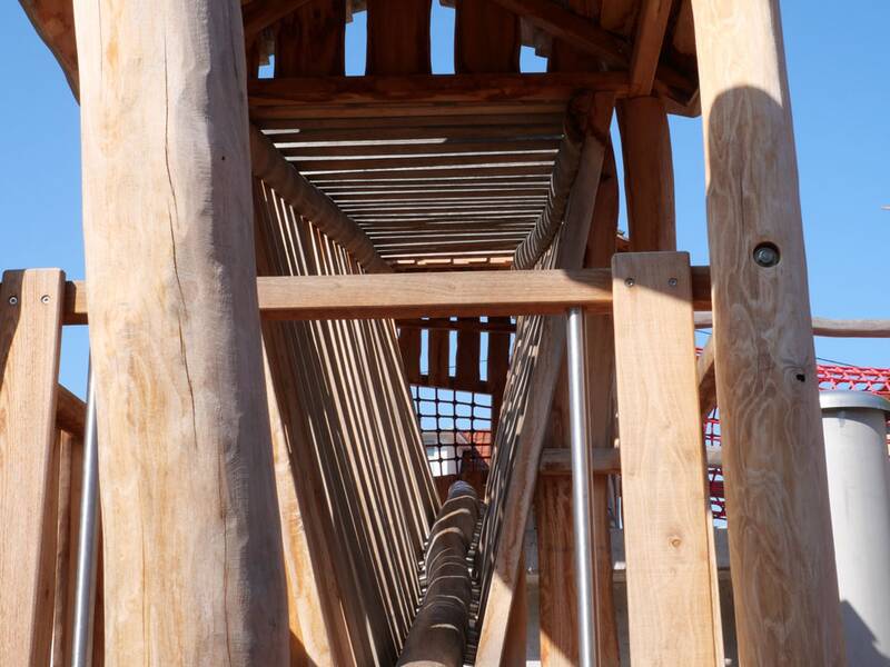 Holzkonstruktion eines Spielturms mit einem geneigten Holzsteg und Netzgeländer.