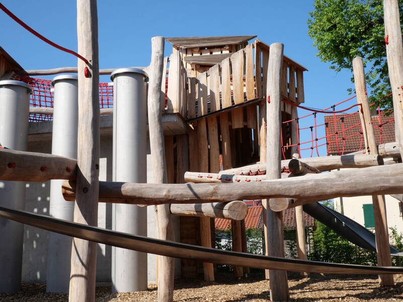 Ein moderner Spielplatz mit Kletterstrukturen aus Holz und roten Seilnetzen steht unter einem klaren blauen Himmel.