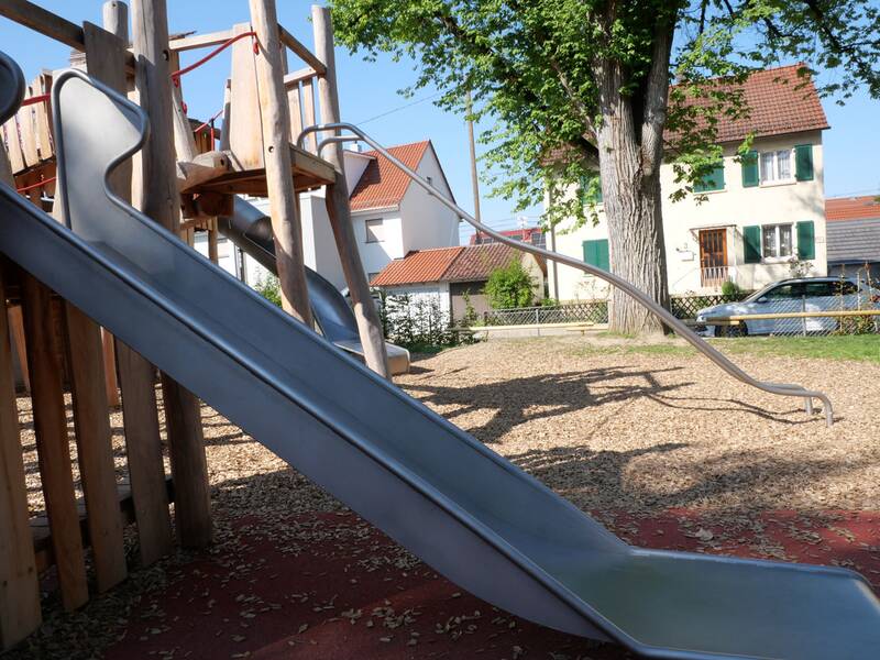 Ein Spielplatz mit mehreren Rutschen und Klettergerüsten aus Holz, umgeben von Bäumen und Wohnhäusern im Hintergrund.