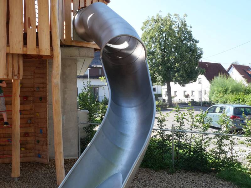 Ein großer Spielturm aus Holz mit einer langen, geschwungenen Metallrutsche steht auf einem Spielplatz, umgeben von Bäumen und einem Wohngebiet im Hintergrund.