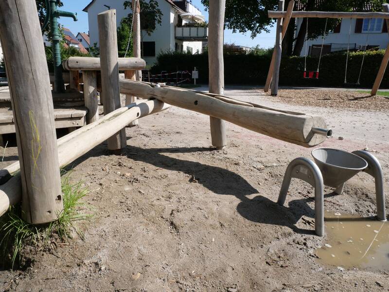 Ein Spielplatz aus Holz mit einer Wasserpumpe und einem Sandbereich, wo Wasser in eine kleine Metallrinne fließt.