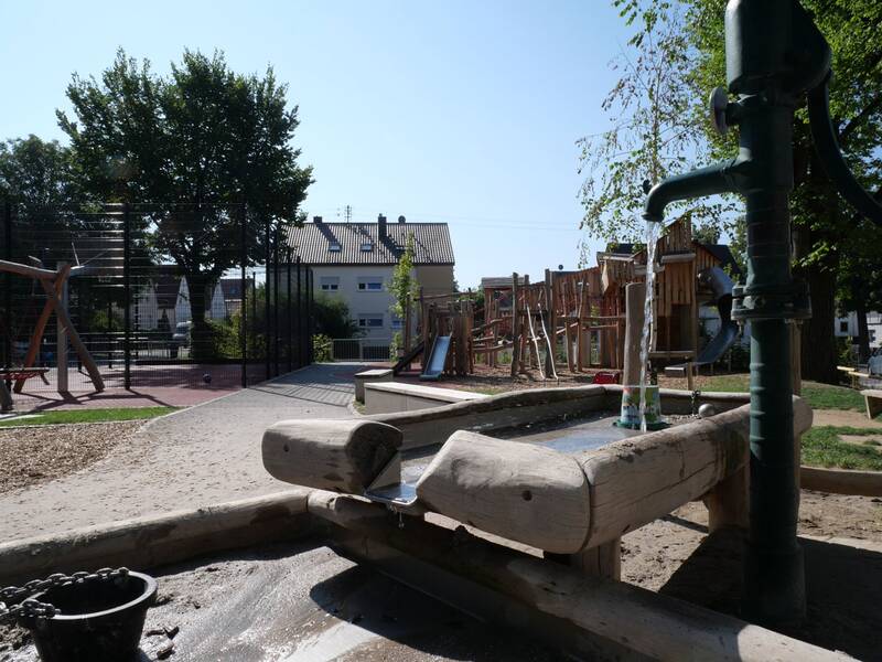 Ein Spielplatz mit einer Wasserpumpe im Vordergrund und verschiedenen Spielgeräten im Hintergrund, umgeben von Bäumen und Gebäuden.