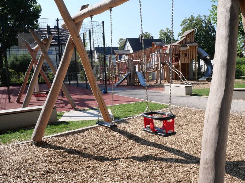 Ein Spielplatz mit Holzklettergerüsten, Rutschen und einer Kleinkindschaukel auf Rindenmulch.