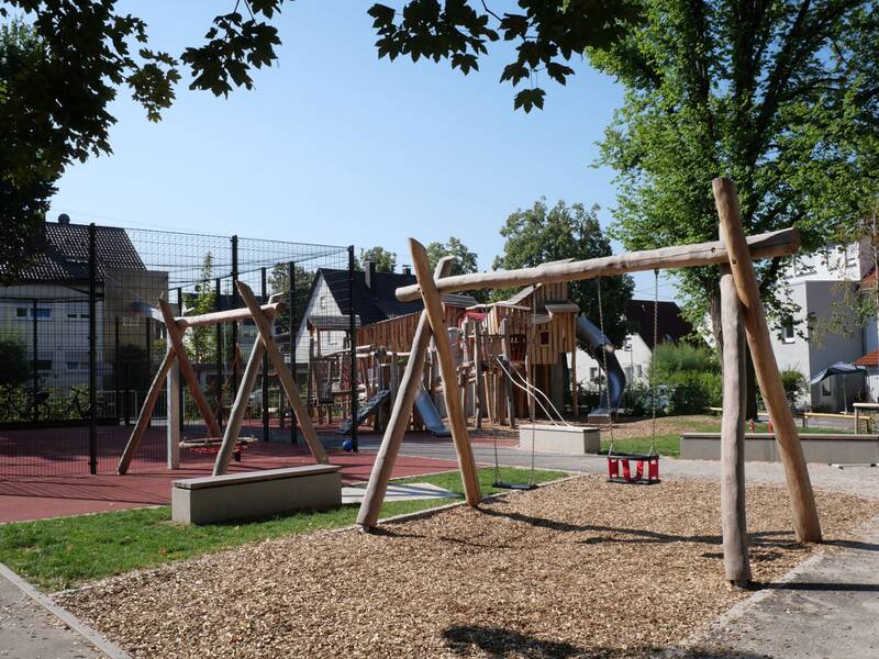Ein Spielplatz mit Holzschaukeln, einem Klettergerüst und einer Rutsche ist umgeben von einem gefüllten Sportplatz und Wohnhäusern im Hintergrund.