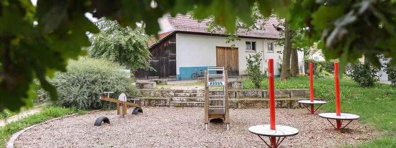 Auf einer von Bäumen umrahmten Fläche stehen eine kleine Rutsche, eine Wippe und drei rote Drehscheiben auf Holzschnitzeluntergrund.