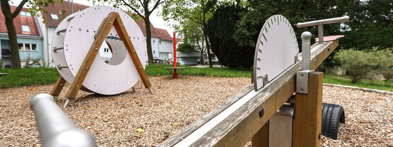 In der Bildmitte steht ein weißes Kletterspielgerät in Form einer Rolle, flankiert von drei runden Drehscheiben mit roten Stangen. Im Hintergrund sind Wohnhäuser und Bäume zu sehen.