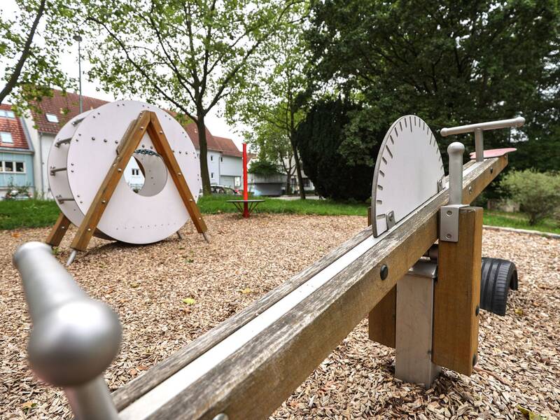 In der Bildmitte steht ein weißes Kletterspielgerät in Form einer Rolle, flankiert von drei runden Drehscheiben mit roten Stangen. Im Hintergrund sind Wohnhäuser und Bäume zu sehen.