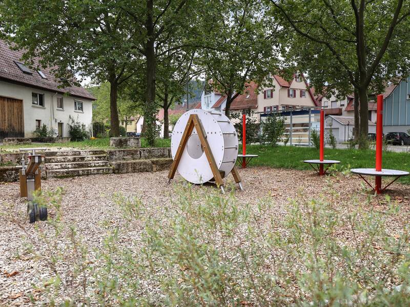 Im Vordergrund ist eine Wippe aus Holz und Metall zu sehen, im Hintergrund ein weißes Kletterelement in Form zweier gegenüberliegender Halbringe.