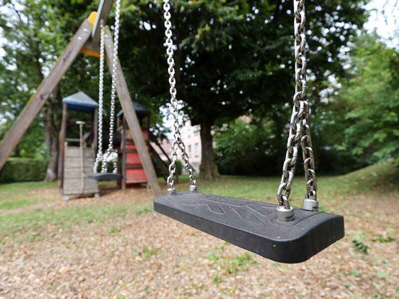 Zwei Schaukeln mit schwarzen Sitzen hängen an einem Holzgestell, im Hintergrund steht ein Spielturm mit Rutsche.