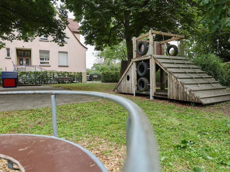 Im Vordergrund ist ein Teil eines Karussells zu erkennen. Dahinter steht ein Holzklettergerüst mit eingelassenen Autoreifen.