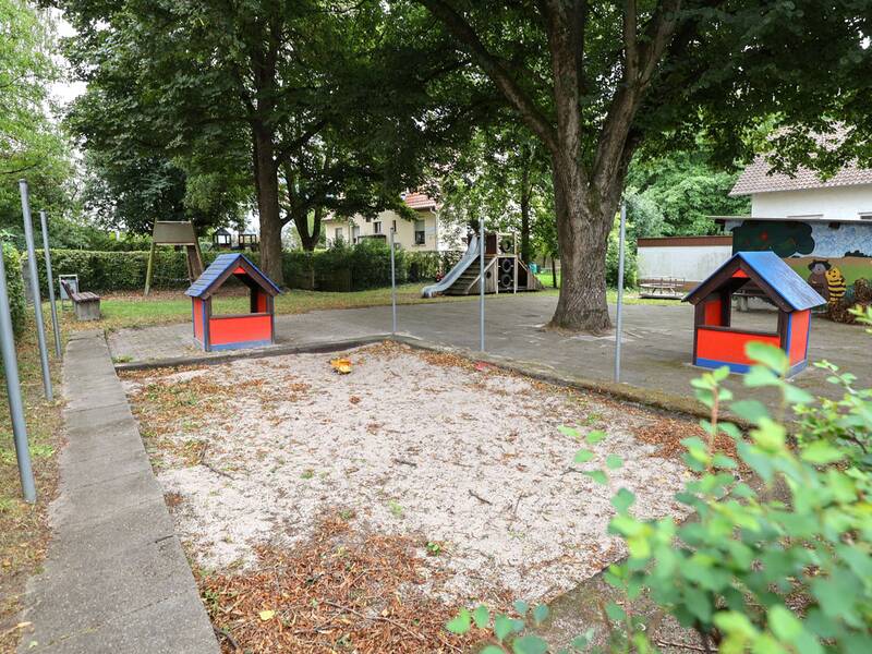 Zwei kleine Spielhäuschen stehen an einem Sandkasten unter Bäumen. Im Hintergrund ist eine Rutsche zu sehen.