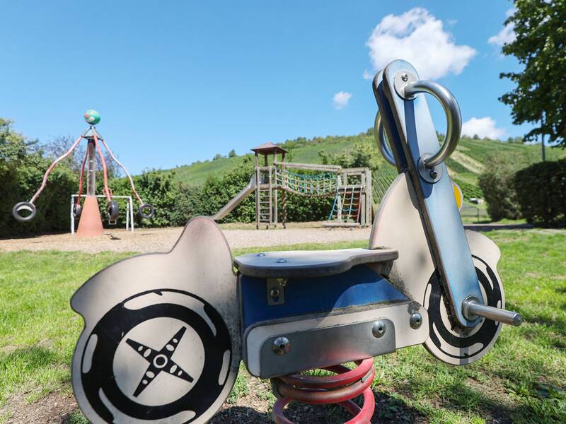 Im Vordergrund steht eine Wippe in Form eines Motorrads, im Hintergrund ein Spielturm mit Rutsche und ein drehbares Kletterkarussell mit Reifen.