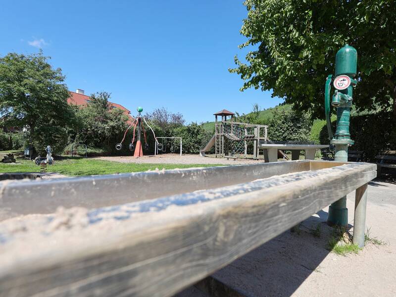 Im Vordergrund eine grüne Wasserpumpe mit Rinne, im Hintergrund Spielgeräte wie Kletterturm, Rutsche und Karussell