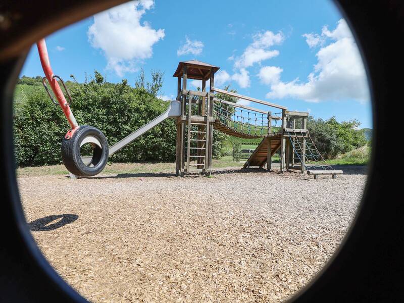 Blick durch eine runde Öffnung auf einen Spielturm mit Rutsche, Kletternetz und einen hängenden Autoreifen.