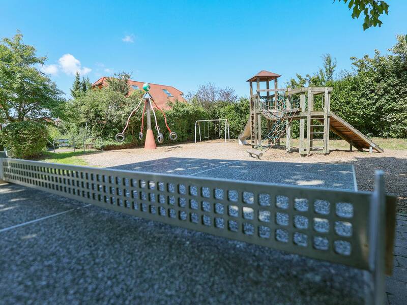 Eine Tischtennisplatte steht vor einem Spielplatz mit Turm, Rutsche, Kletternetzen und einem drehbaren Karussell.