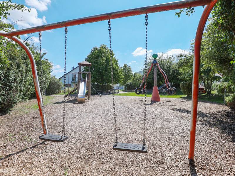 Zwei Schaukeln hängen an einem roten Metallgestell, dahinter ein Spielturm mit Rutsche und ein Karussell mit Reifen.