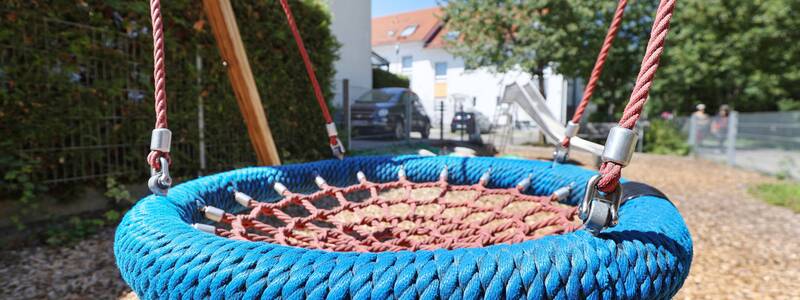 Eine Nestschaukel mit blauem Rand hängt an roten Seilen, im Hintergrund sind Häuser und eine Rutsche zu sehen.