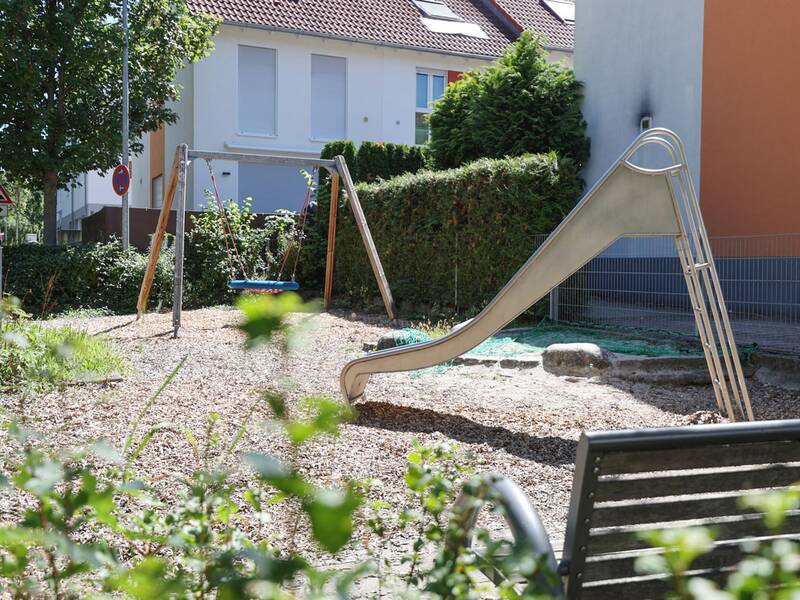 Eine Nestschaukel mit blauem Rand hängt an roten Seilen, im Hintergrund sind Häuser und eine Rutsche zu sehen.