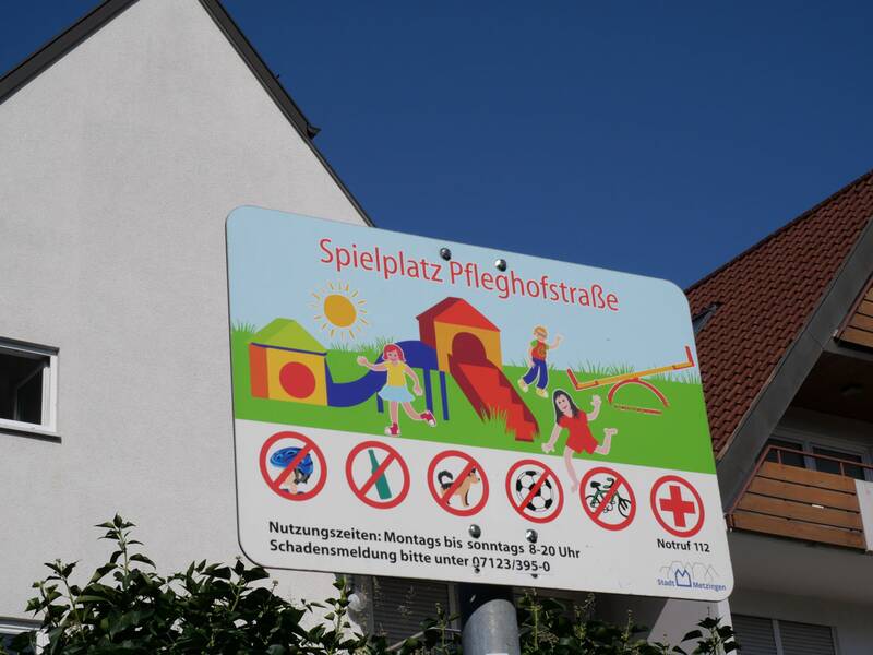 Ein Schild mit der Aufschrift „Spielplatz Pfleghofstraße“ zeigt bunte Zeichnungen spielender Kinder und Symbole mit Verboten.