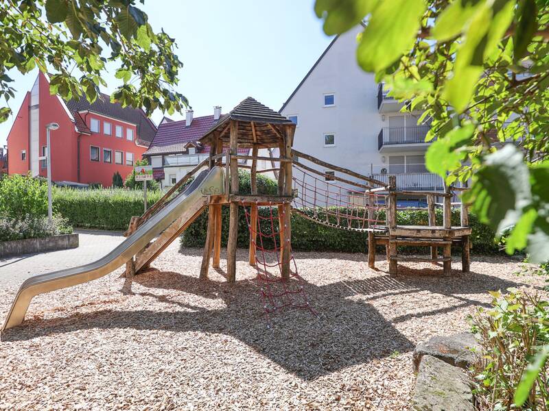 Ein Spielplatz mit Holzkletterturm, Edelstahlrutsche und Kletternetzen auf einem mit Rindenmulch bedeckten Boden.