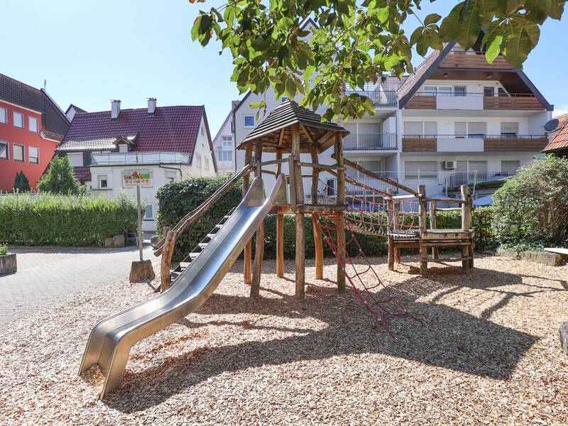 Ein Spielplatz mit Holzkletterturm, sichtbar sind Kletterelemente aus Holz und Netz sowie umliegende Wohnhäuser.