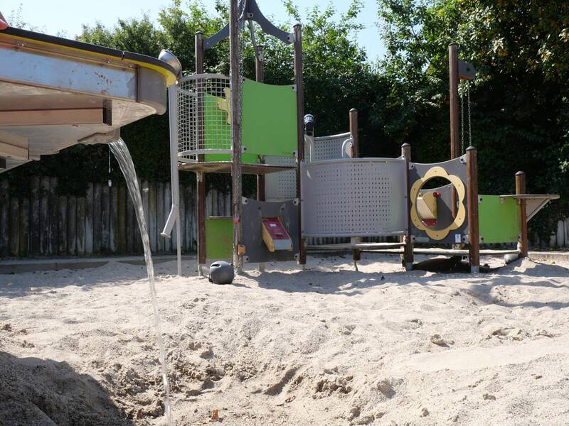 Ein Wasserstrahl fließt aus einem Metalltisch in einen Sandbereich, im Hintergrund steht ein buntes Kletter- und Spielgerät mit grünen und grauen Flächen.