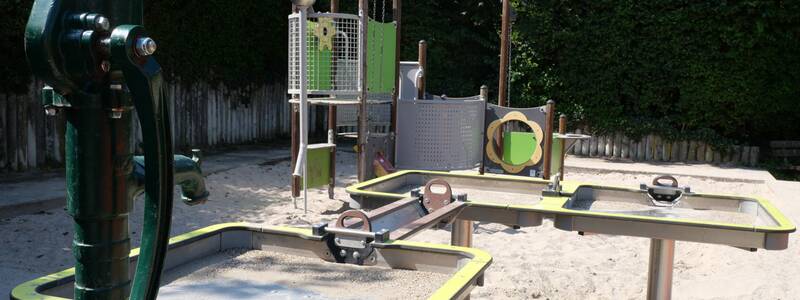 Ein Wasserspieltisch. Im Vordergrund ist eine grüne Wasserpumpe zu sehen, im Hintergrund ein Sandspielhaus.