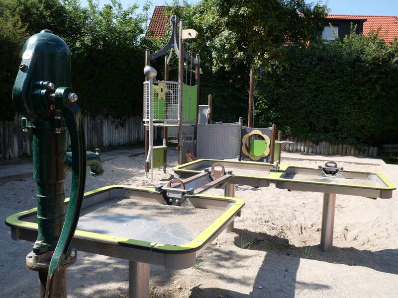 Ein Wasserspieltisch. Im Vordergrund ist eine grüne Wasserpumpe zu sehen, im Hintergrund ein Sandspielhaus.