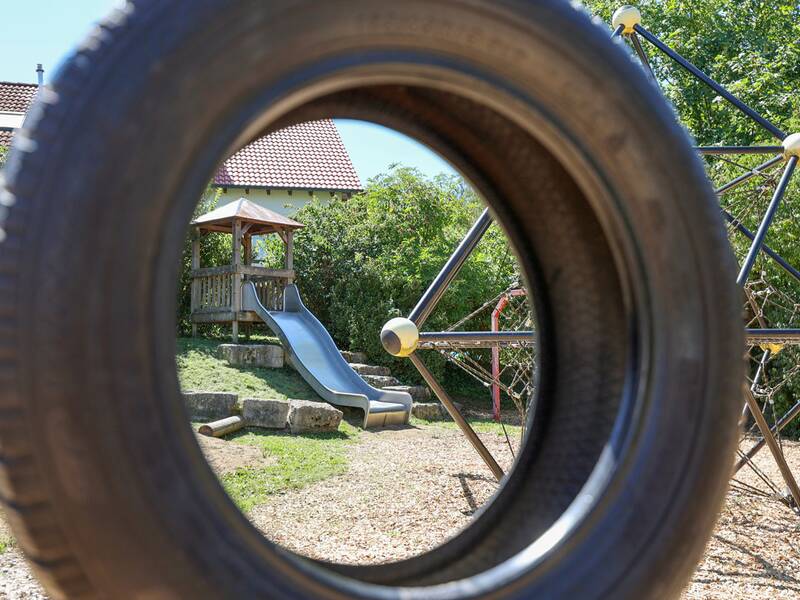 Eine Tischtennisplatte steht auf einem gepflasterten Bereich neben einem Spielplatz mit Wippe, Kletternetz und weiteren Geräten.
