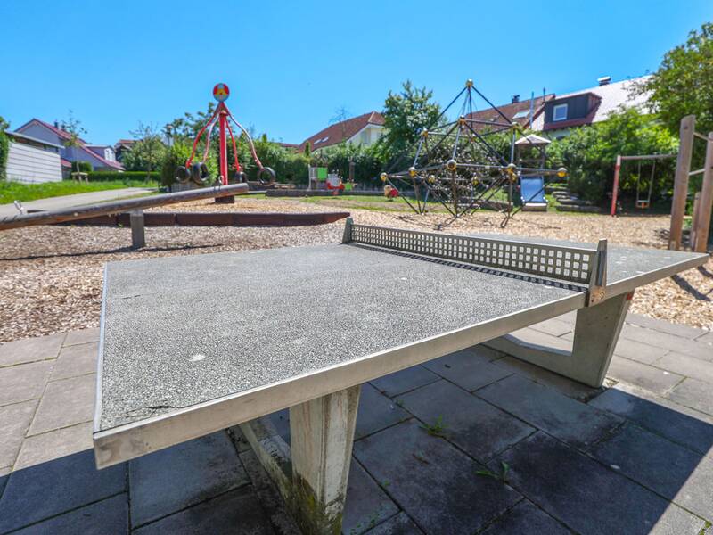 Eine Tischtennisplatte steht auf einem gepflasterten Bereich neben einem Spielplatz mit Wippe, Kletternetz und weiteren Geräten.