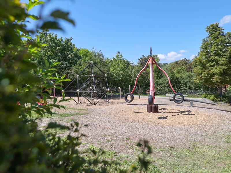 Ein Spielplatz mit großem Kletternetz, drehbarem Reifengerät und Bäumen im Hintergrund bei sonnigem Wetter.