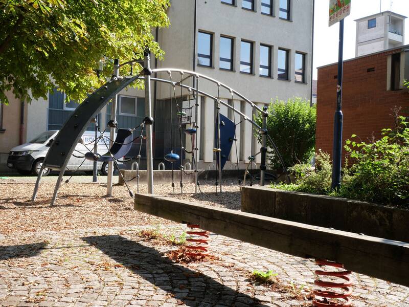 Ein Kletterbogen, im Vordergrund eine schmale Wippe auf roten Federn. Links parkt ein weißer Transporter, im Hintergrund stehen Büro- und Backsteingebäude.