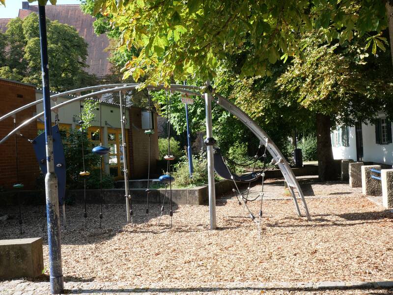 Ein moderner silberner Kletterbogen mit Hänge- und Balancierelementen steht auf einem Platz mit Holzschnitzeln. Im Hintergrund sind Bäume, ein Backsteingebäude und gelbe Türen zu sehen.