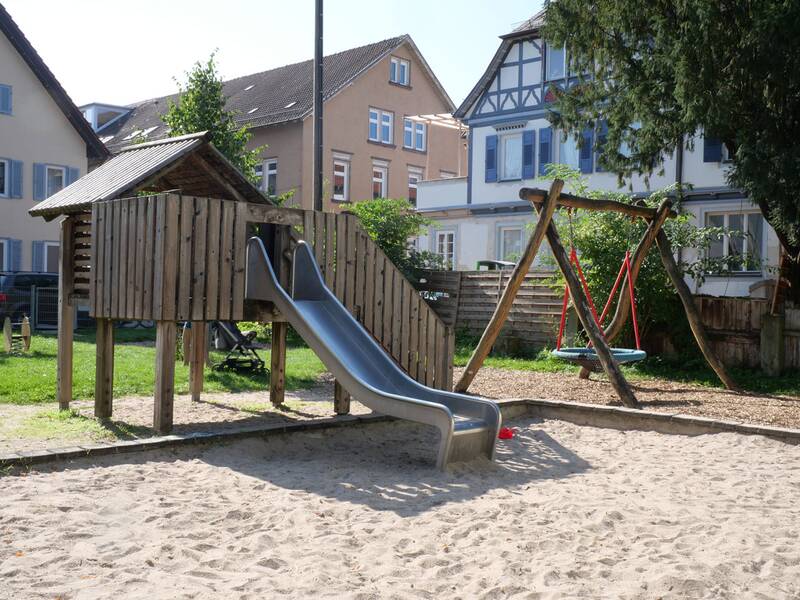 Ein Holzhaus mit Dach und Rutsche steht auf einem Sandbereich, daneben eine Nestschaukel vor Wohnhäusern.
