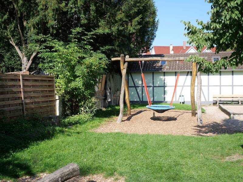 Ein Holzgestell mit einer runden Nestschaukel steht auf einer Rasenfläche, dahinter ein kleines Fachwerkgebäude.